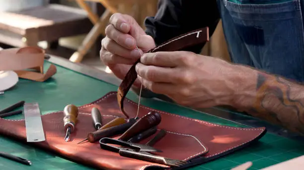 Close-up hands handmade leathermakers brown leather bag strap handle made animal skin other hand holding needle needle is being used remove thread untie bag strap, as a solution.
