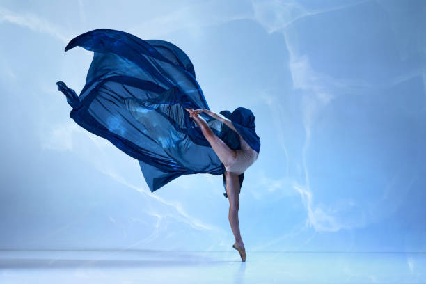 istock Young classical dancer dancing on fingertips with silk fabric over blue background. Beauty and grace in movements 1477521706