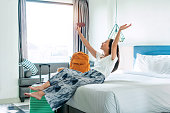 Happy young Asian traveler woman relax on bed in hotel room. Travel alone, summer and holiday concept.