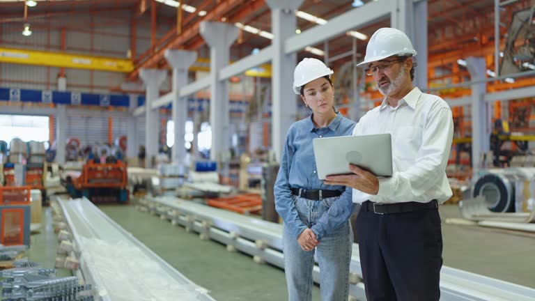 Two professional engineer man and woman manager leader wearing helmet and holding laptop standing in factory talking and discussing for work