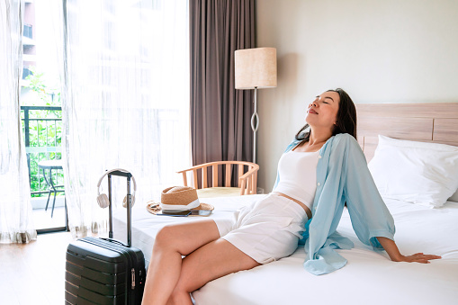 Carefree young Asian traveler woman relax on bed in hotel room. Travel alone, summer and vacation concept. Copy space