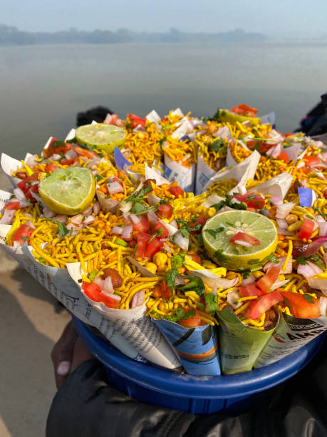 Stock photo showing close-up, elevated view of display of paper cones filled with chaat of savoury salted snacks (namkeen) for sale in blue, plastic bucket. imagen de una persona irreconocible sosteniendo un cubo de plástico azul que muestra conos de papel llenos de chaat (bocadillos salados), namkeen y bhel puri (arroz inflado y verduras), bocadillos salados salados para la venta, vista elevada, enfoque en p