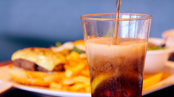 Close-up of pouring brown soft drink with ice cubes and lemon into glass, set against fast food burger and fries background. Unhealthy food concept.