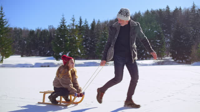 Slow motion wide tracking shot of a senior man pulling his young granddaughter on a sled across a frozen lake in sunshine and having fun. Shot in Slovenia.