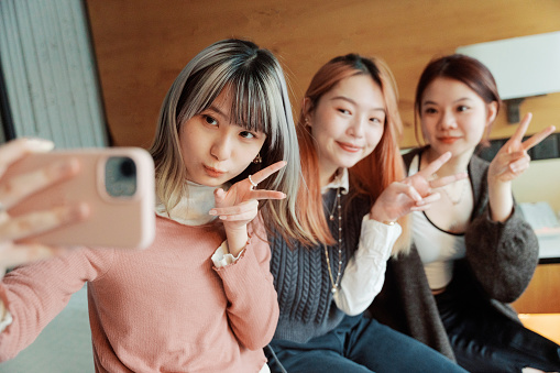 Side medium shot of three smiling young Asian women sitting on a bed, holding and using a smartphone or mobile phone, taking a selfie picture or photo together while posing cutely in front of the bright window, to create memory for their trip or celebrate friendship.\n\nYoung happy Asian female friends besties, sisters or female travelers or tourists, having fun relaxing in a luxurious western hotel room on a holiday or vacation.
