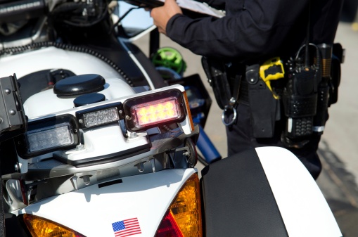 The rear end of a police motorcycle with an officer standing to the side slightly blurred on purpose as he writes a ticket.