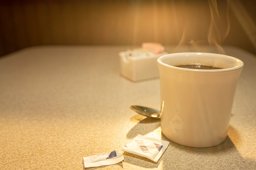 A lone steaming hot cup of coffee with steam rising, along with an open sugar packet, as the light from the sun rises through the window of a diner.