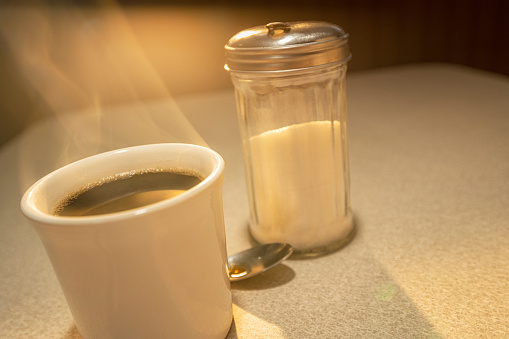 A lone steaming hot cup of coffee with steam rising, along with a sugar dispenser, casting a long shadow as the light from the sun rises through the window of a diner.