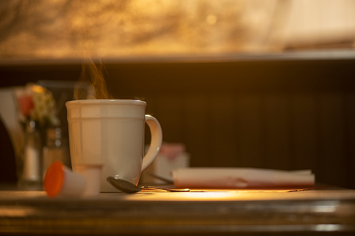 A close-up of the top of a steaming hot mug coffee with steam rising as the light from the sun rises through the window of a diner.