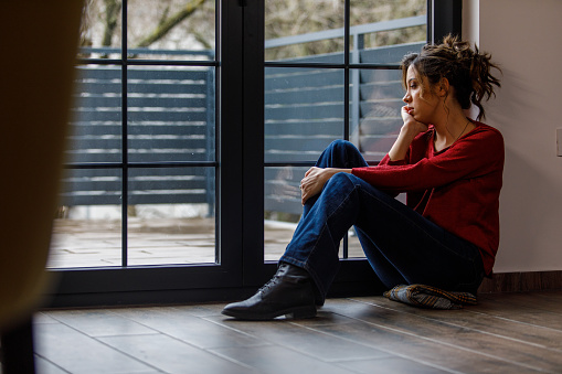 Wide shot of anxious young woman sitting on the floor, by the window, hugging self and trying to calm down while struggling with depression and negative thoughts. She is looking outside, contemplating.