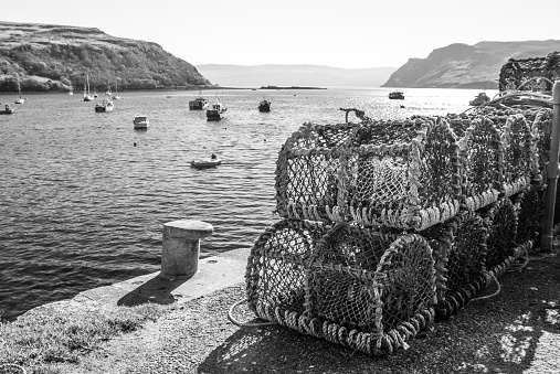 Stack of crab traps with the bay of Portree in the background, in black and white. Portree is a fishing village overlooking a sheltered bay on the east side of the Isle of Skye. Although this village is only about 200 years old, it is the largest village on the Island and the Islands Capital.