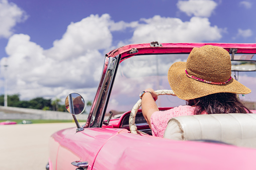 Anonymous female traveler in straw hat driving pink retro cabriolet on cloudy day during summer vacation in Cuba