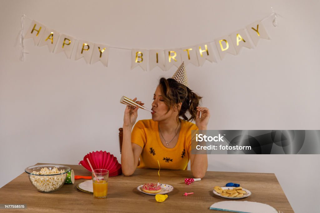 Cheerful young woman blowing a party horn blower during a solo birthday celebration Front view of joyful young woman sitting at table, wearing a party hat and blowing a party horn blower during a solo birthday celebration. ''Happy birthday'' banner hanging on the wall behind her. 20-24 Years Stock Photo