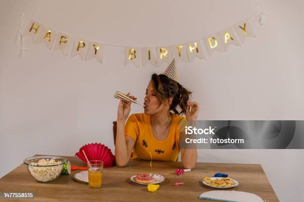 Cheerful Young Woman Blowing A Party Horn Blower During A Solo Birthday Celebration Stock Photo - Download Image Now