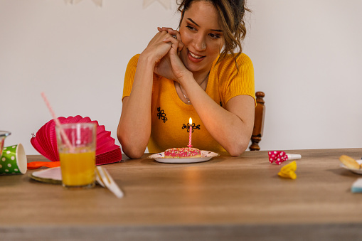 Front view of happy young woman sitting at table, hands clasped by her face, smiling, looking down and making a wish before blowing a birthday candle in a doughnut while celebrating her birthday alone.