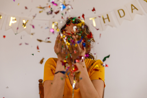 Front view of joyful young woman sitting at table during a solo celebratory party, looking at camera and blowing handful of colorful confetti. ''Happy birthday'' banner hanging on the wall behind her.