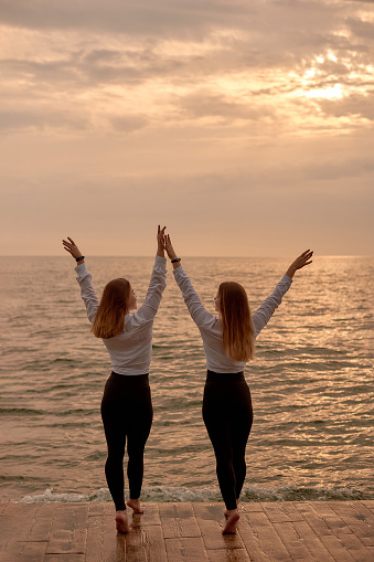 Healthy start meet sunrise - two sisters twin girls in identical clothes on sea background. dancing couple two woman twins training in morning on beach.