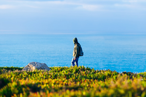a tourist in a green sweater with a hood walking on the picturesque shore of the ocean