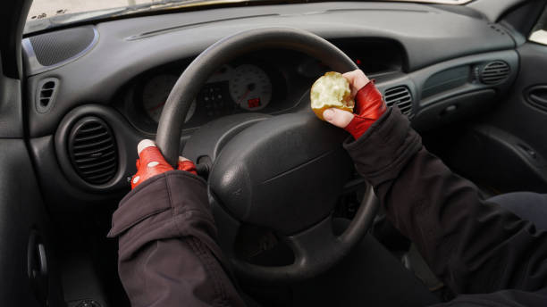 eating while driving hands on the wheel of a car in red gloves, with an apple in one hand a person drives a car. Concept: Eating while driving a car prison meal stock pictures, royalty-free photos & images