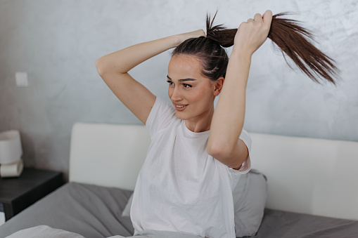 This stock photo depicts a young woman sitting cross-legged in her bed, using her fingers to tie her hair back into a loose bun. She's wearing a white t-shirt and has a contented expression on her face