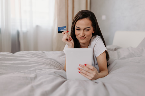 A young woman is seen laying on her bed, holding a digital tablet and a credit card, as she browses online stores and makes purchases