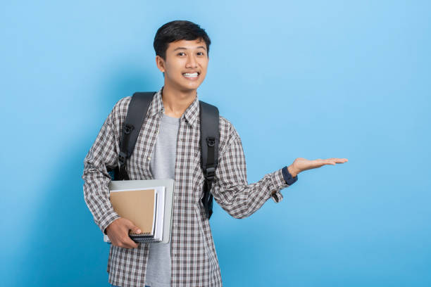 istock Young asian college student carrying backpack showing present product and advertising with hands isolated over blue background 1477580276