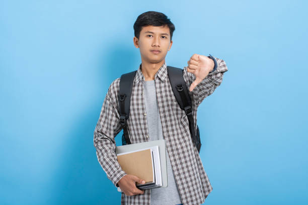 istock Young asian college student carrying books and backpack expressing negative doing thumbs down gesture isolated over blue background 1477580553