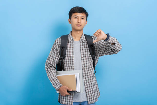 istock Young asian college student carrying books and backpack expressing negative doing thumbs down gesture isolated over blue background 1477580606