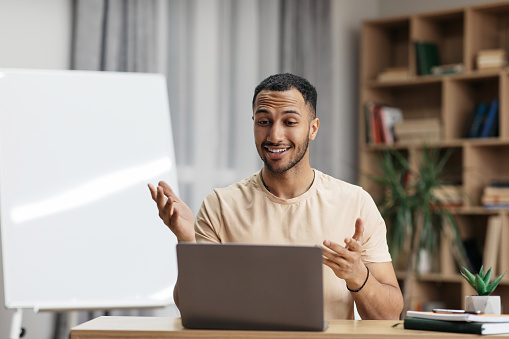 Cheerful arab male teacher looking at laptop and gesturing, sitting at desk near blackboard at home interior. Education, study and tutoring, new normal