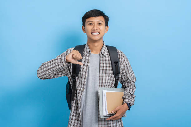 istock Young asian college student carrying books and backpack self confident pointing finger at himself isolated over blue background 1477582643