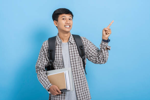 istock Young asian college student carrying books and backpack pointing with finger on blank copyspace isolated over blue background 1477583024