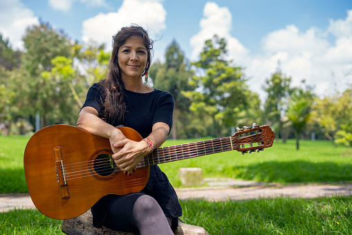 Beautiful Latin woman holds a guitar sitting on a wooden log outdoors in the park.