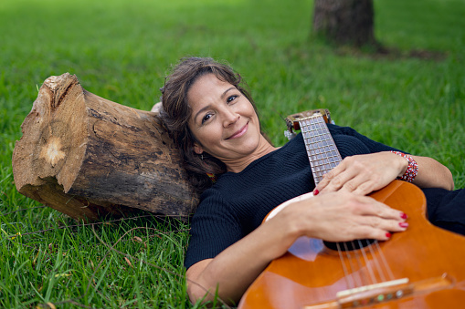 Portrait of Beautiful Latin woman looking at the camera holds a guitar lying on a wooden log outdoors in the park.