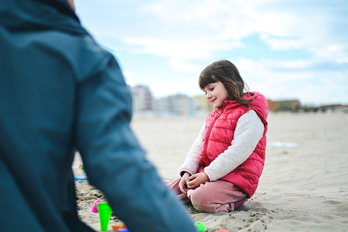 daughter in a warm clothing sitting on a sand and playing with father.