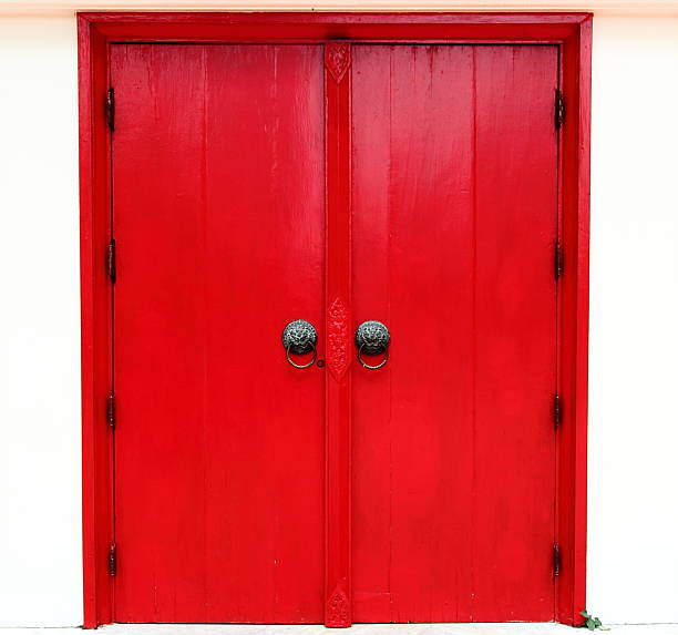 Red wooden entrance door in a tenement in Kalisz, with round glass windows, concrete facade, and yellow installation sign on the wall.
