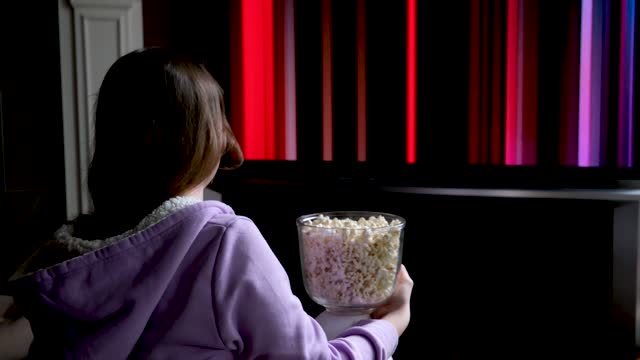 teenage girl sits on floor and watches netflix on big screen of TV screensaver slow motion flashes in hands of young woman popcorn girl takes it and eats it spellbound watching expectation of movie