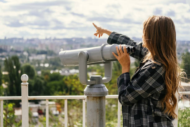 Curious young woman is pointing at the city while holding hand on binoculars. stock photo
