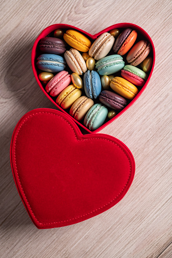 Round baked multicolored macarons lie in a red heart shaped box on wooden background