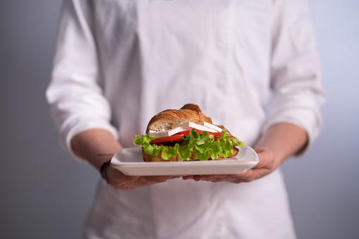 Chef holding a plate with a tasty croissant sandwich on gray background.