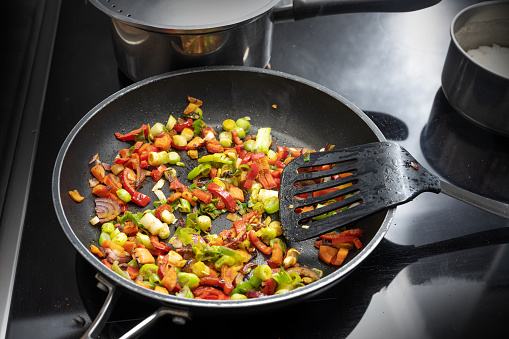 Stir fried vegetables like bell pepper, onion and leek with spices in a black frying pan on the stovetop with other pots, cooking a vegetarian curry, copy space, selected focus, narrow depth of field