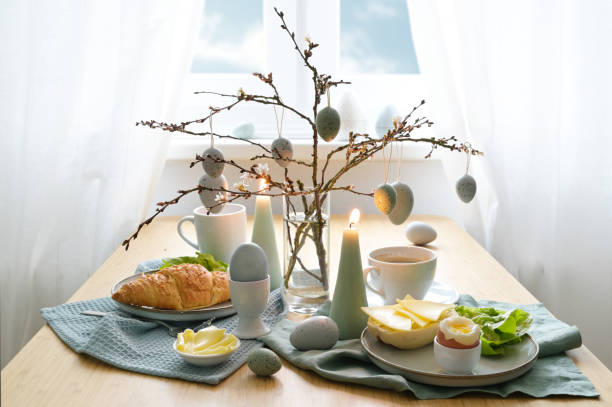 istock Breakfast table with hanging Easter eggs on spring branches, candles, and blue gray decoration in front of a window with white curtains, copy space, selected focus 1477619215