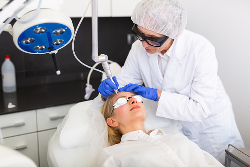 Female cosmetologist making laser mole removal procedure for young woman.