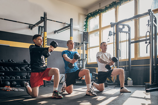 Three people, fitness instructor training with two teenage boys with dumbbells in gym.