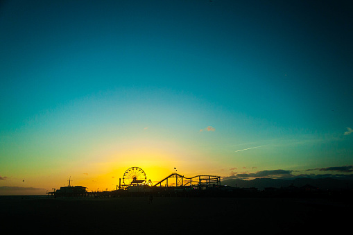 The sun setting behind Santa Monica Pier, with a Ferris wheel and roller coaster at Pacific Park in silhouette.