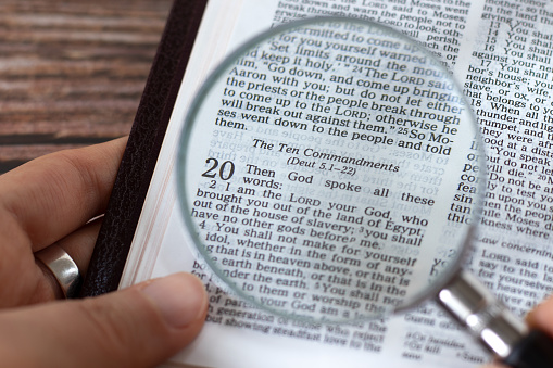 Human hand holding a magnifying glass over open holy bible book of Exodus verses for Ten Commandments. Top view. A close-up. Studying God's righteous spiritual Law, biblical concept.
