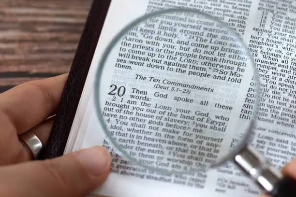 Photo of Human hand holding a magnifying glass over open holy bible book of Exodus verses for Ten Commandments, top view