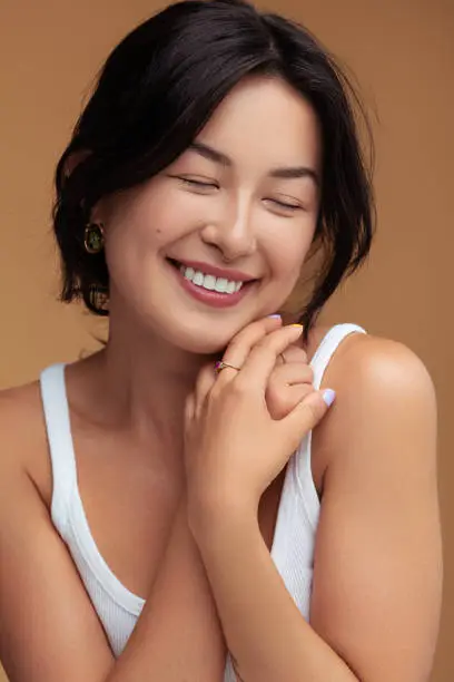 Timid young Asian female clasping hands on shoulder and smiling with closed eyes against brown background