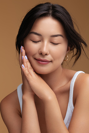 Positive Asian female with dark hair clasping hands near cheek and closing eyes during skin care routine against brown background