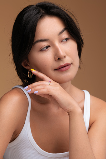 Young Asian female in white top with dark hair looking away and touching soft skin of cheek during beauty routine against brown background