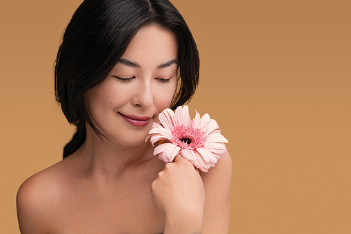 Beautiful young ethnic female model with pure skin performing perfect cosmetic skincare holding blooming flower on brown background
